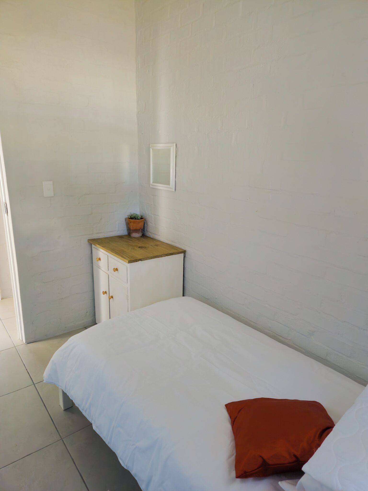 A small, clean room with a single bed, a nightstand with a plant, and a white wall.