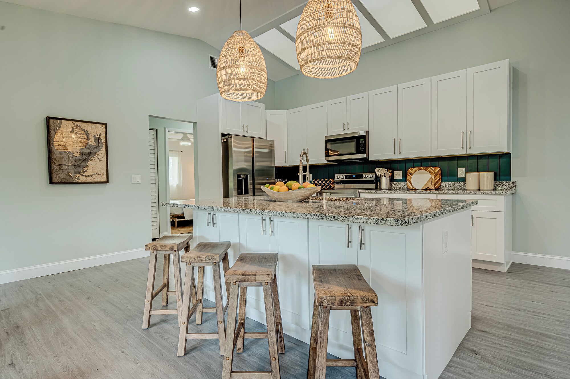 A spacious kitchen with white cabinets, a large island with stools, a hanging light fixture, and a view into the adjacent living area.