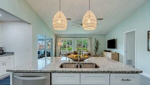 A modern kitchen with a granite countertop, white cabinetry, and a built-in dishwasher. There are two pendant lights hanging above the island, and a large window provides a view of the outdoors. - Gorgeous Home with Heated Pool Near Beaches (Seminole)