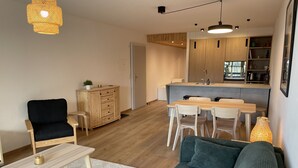 Een moderne woonkamer met een bank, een salontafel en een eethoek met een tafel en stoelen. Er is een keuken met houten keukenkastjes en een spoelbak.
