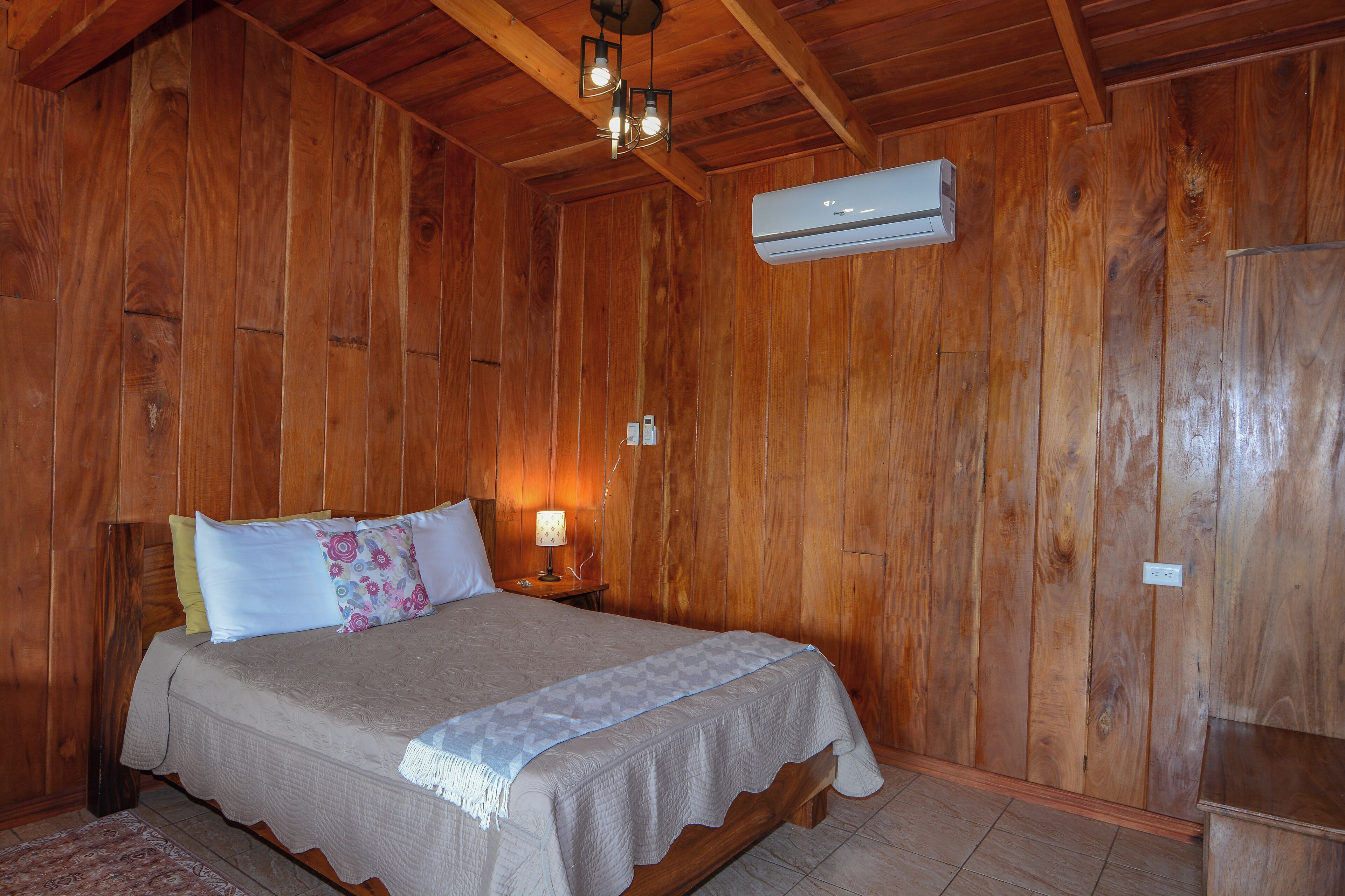 A wooden-paneled room with a bed, a nightstand, an air conditioning unit, and a lamp.