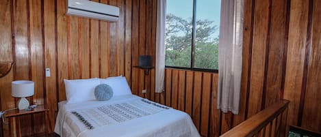 A wooden room with a bed, a nightstand, a lamp, and a window with curtains.