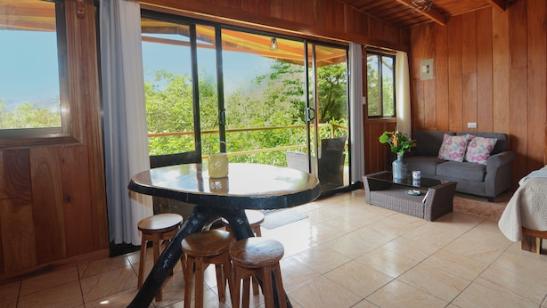 A wooden interior with a dining table, stools, a sofa, and a large window offering a view of greenery.
