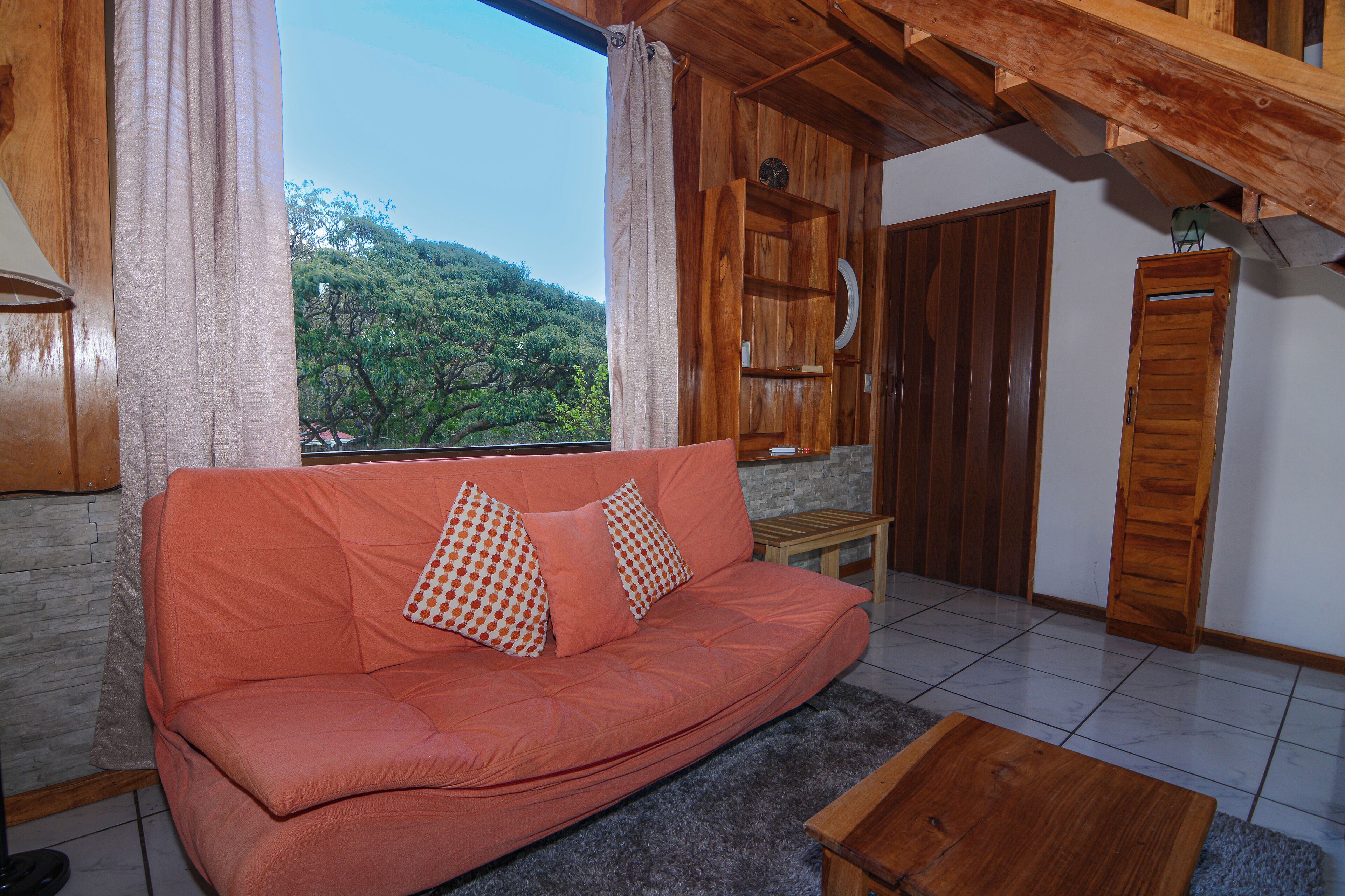 A cozy living room with a sofa, a coffee table, and a view of a forested area through a window.