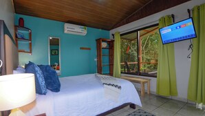 A bedroom with a bed, a TV, a window with curtains, a mirror, and a shelf with decorative items.