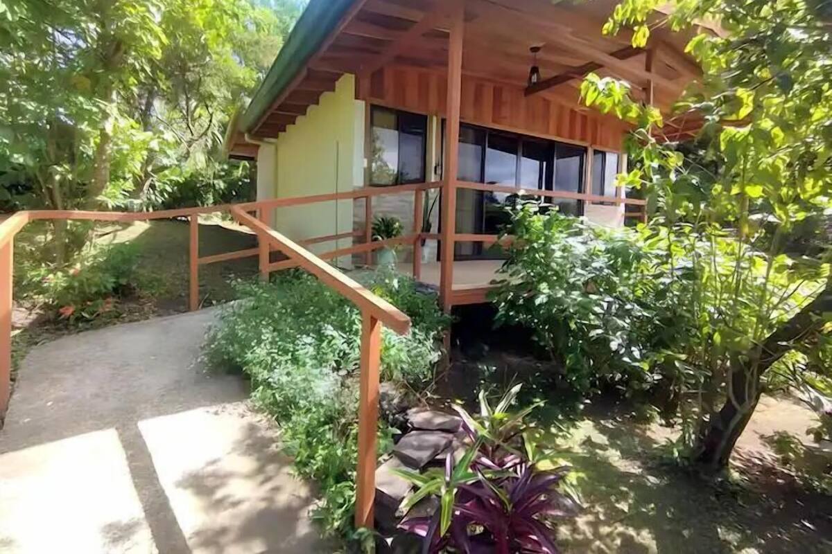 Una casa de fusta amb un porxo, envoltada de vegetació exuberant i un jardí petit.