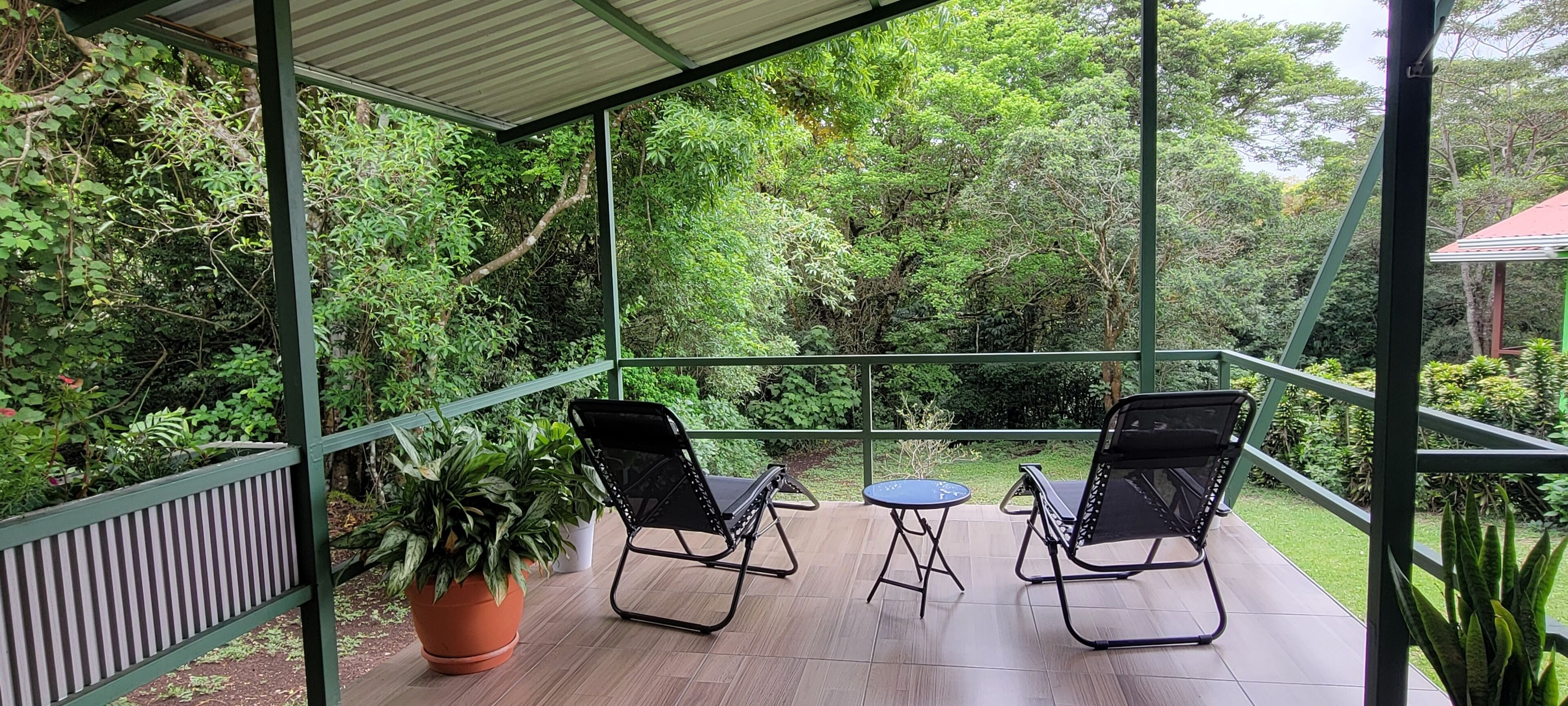 A covered outdoor area with two chairs and a table, surrounded by greenery.