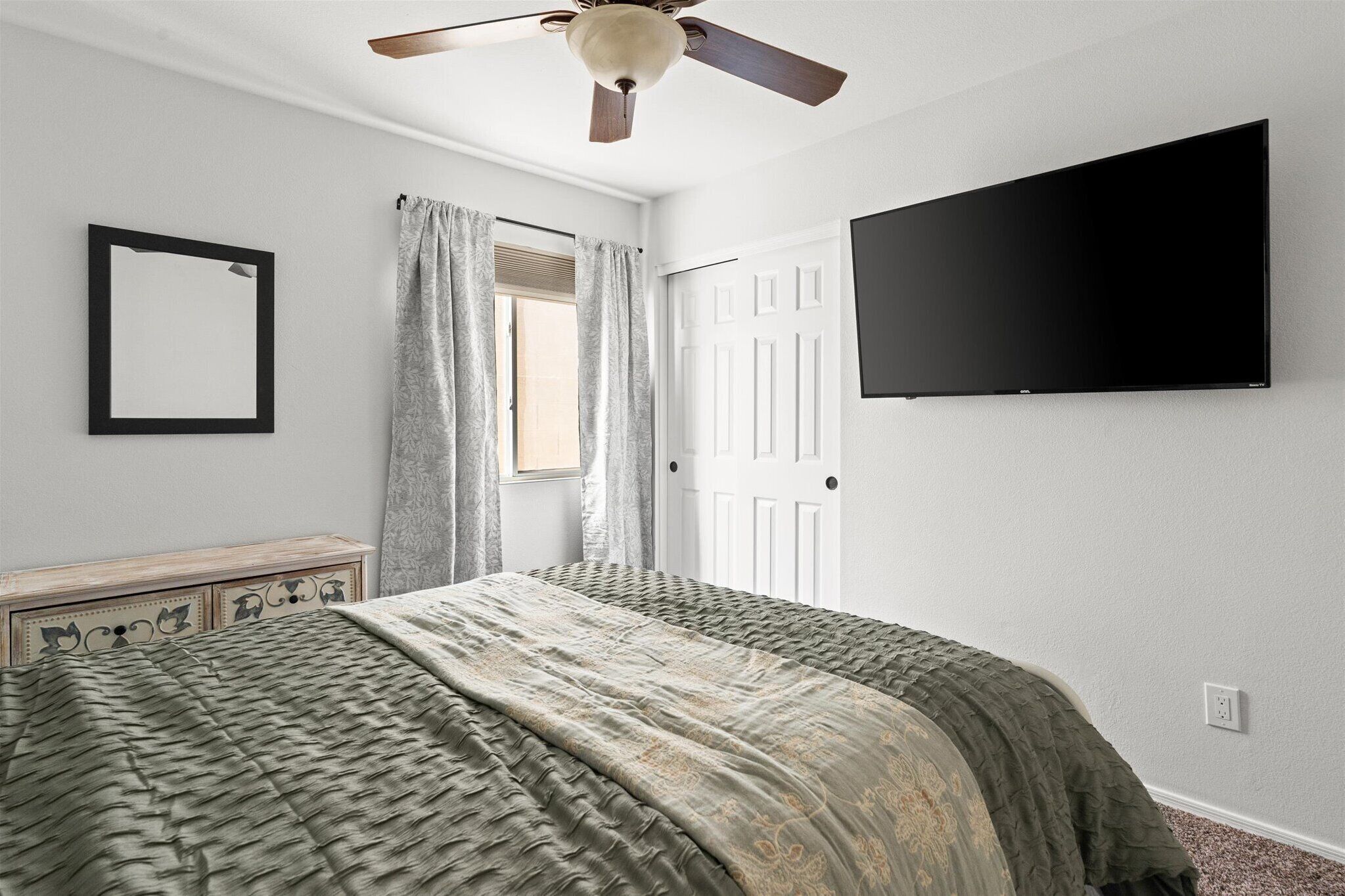 Une chambre à coucher avec un lit, un ventilateur de plafond, un téléviseur fixé au mur et une fenêtre avec des rideaux.