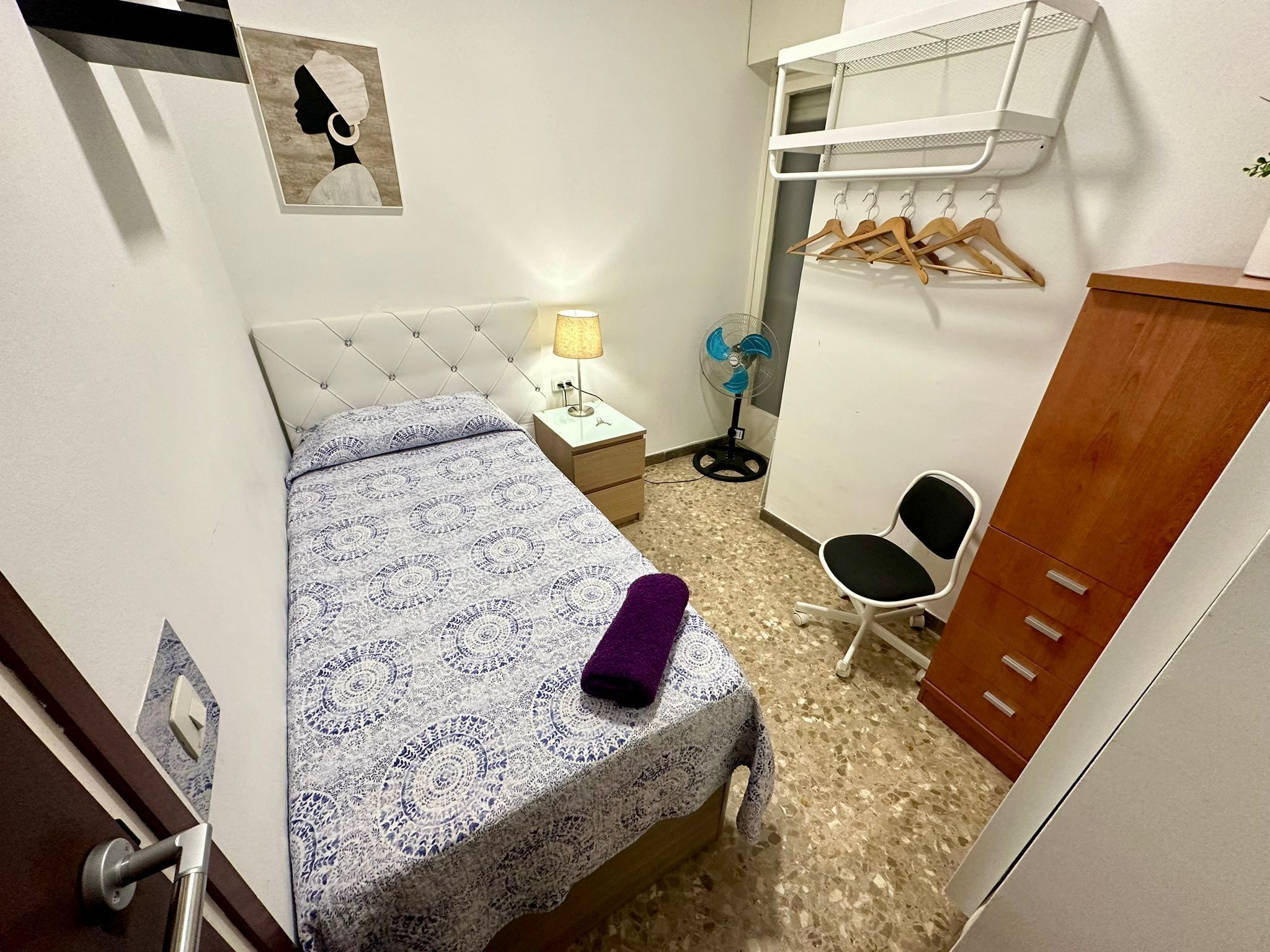Un dormitorio pequeño con cama, mesita de noche, lámpara, ventilador, silla y armario.