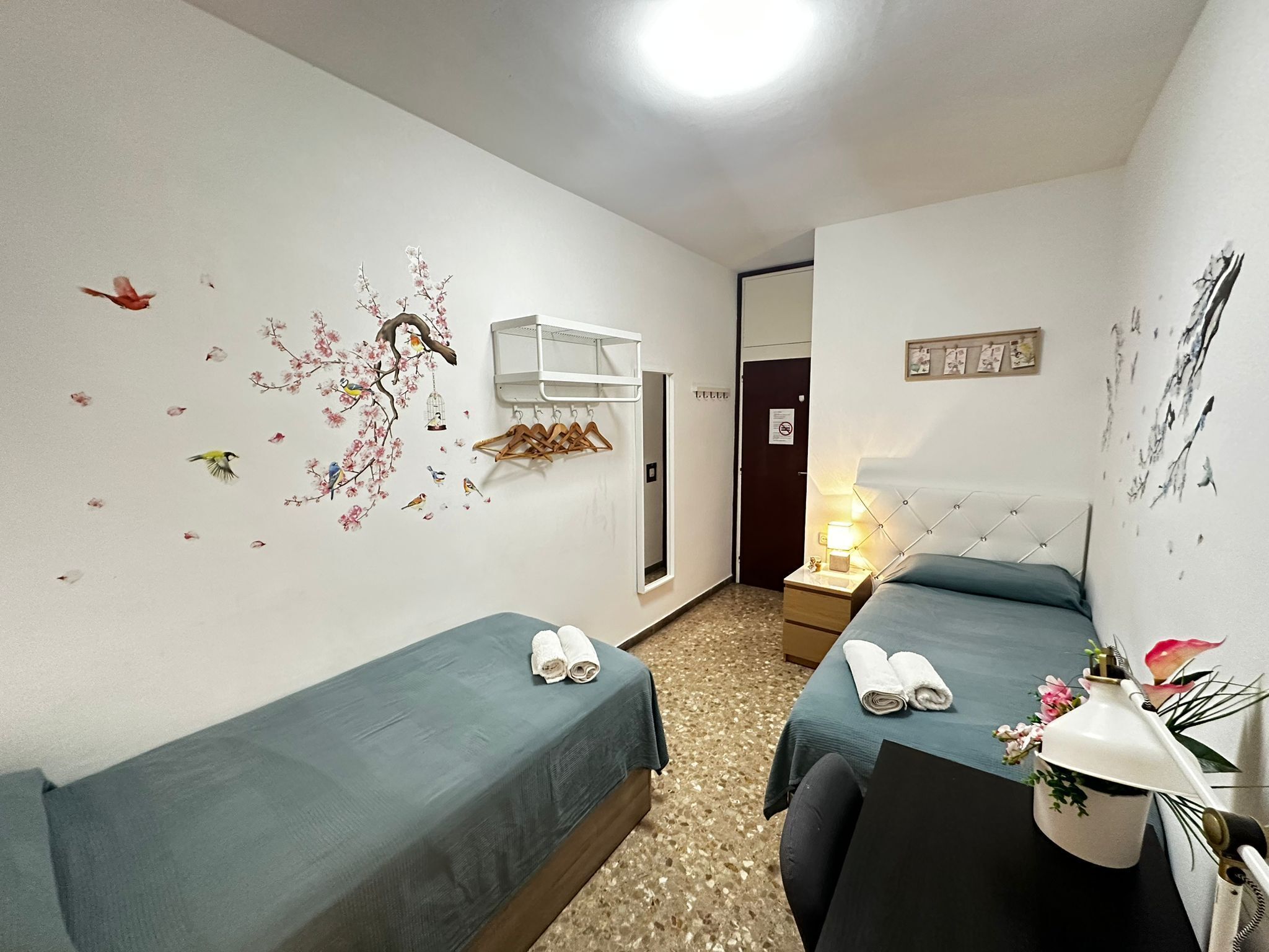 A hotel room with two single beds, a nightstand with a lamp, a wall-mounted drying rack, and a door with a 'No Smoking' sign.