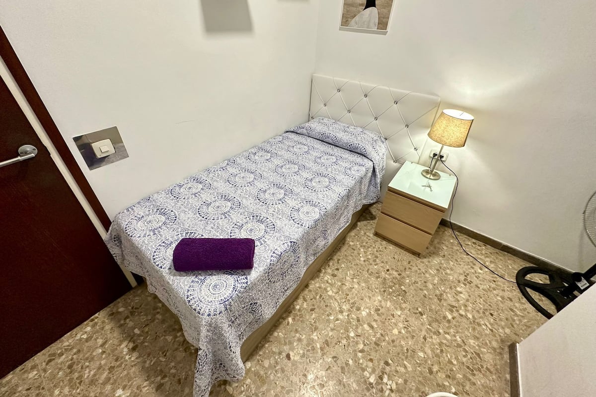 Un letto piccolo e ordinato con una coperta a fantasia e un asciugamano viola, un comodino con una lampada, e un'opera d'arte appesa al muro.