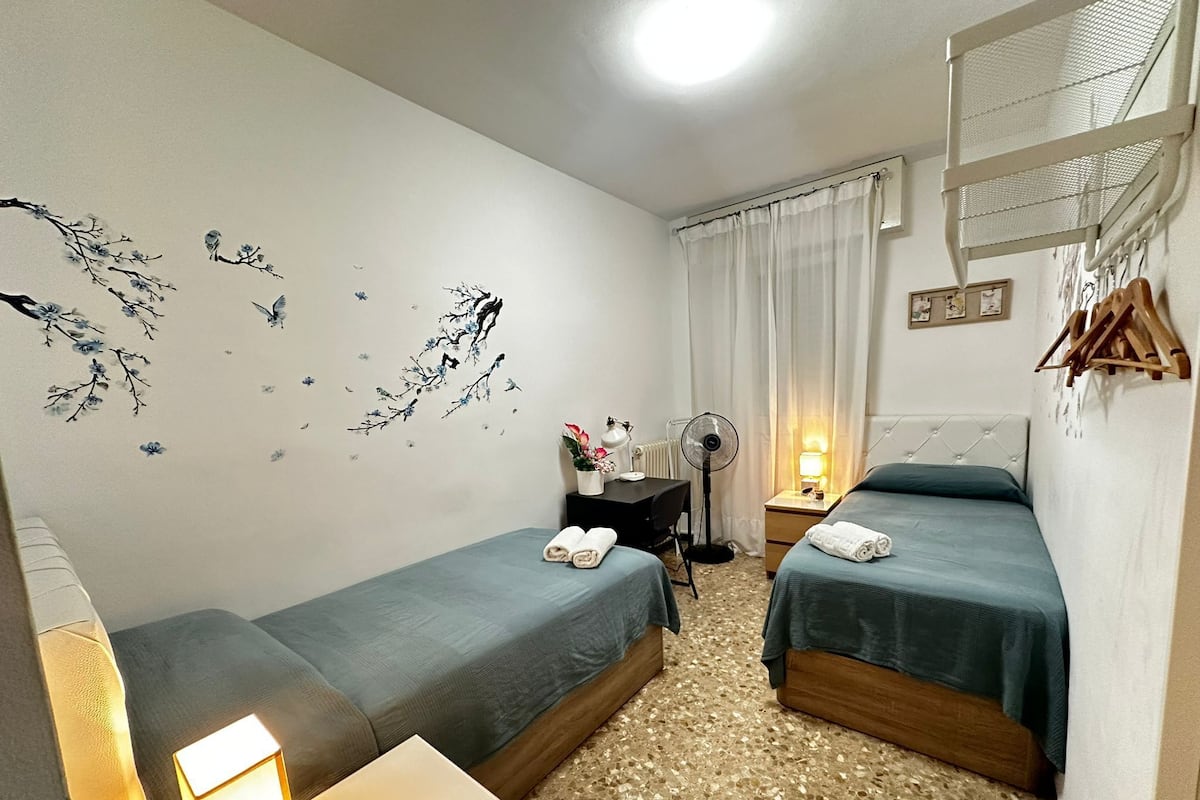 Camera d'albergo con due letti singoli, una scrivania con una sedia, una ventola da terra, un tavolino con una lampada e un'asciuga-fiori a parete.