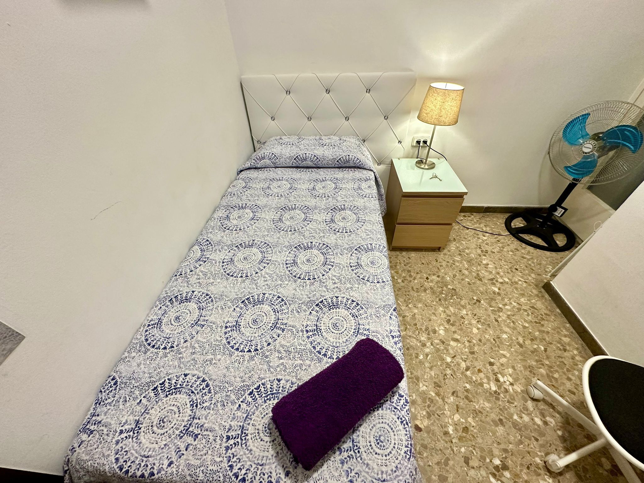 A small, neatly made bed with a patterned bedspread and a purple throw, a bedside table with a lamp, a standing fan, and a chair with wheels.