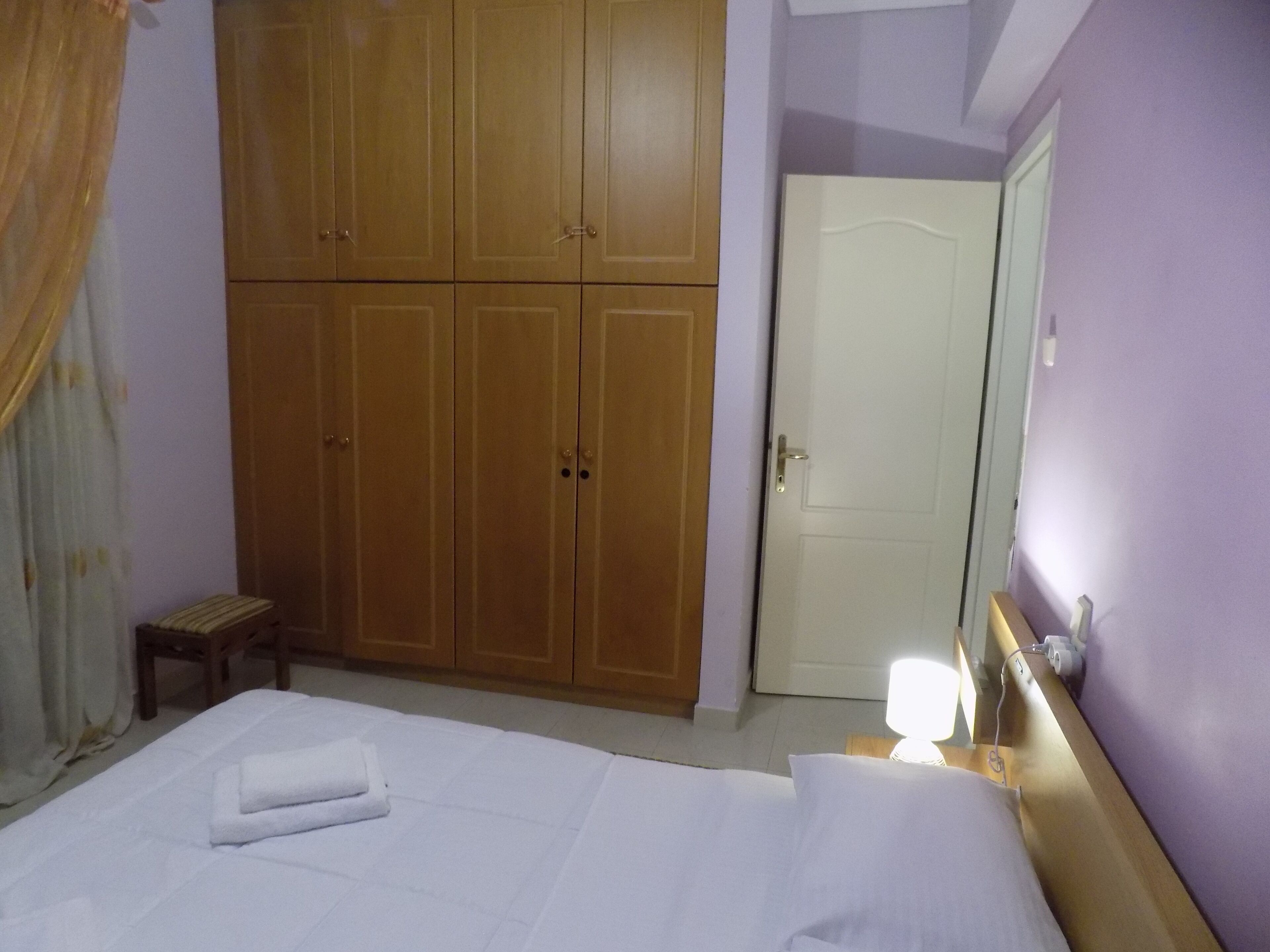 A hotel room with a large wardrobe, a bed with white linens, a bedside table with a lamp, and a door.