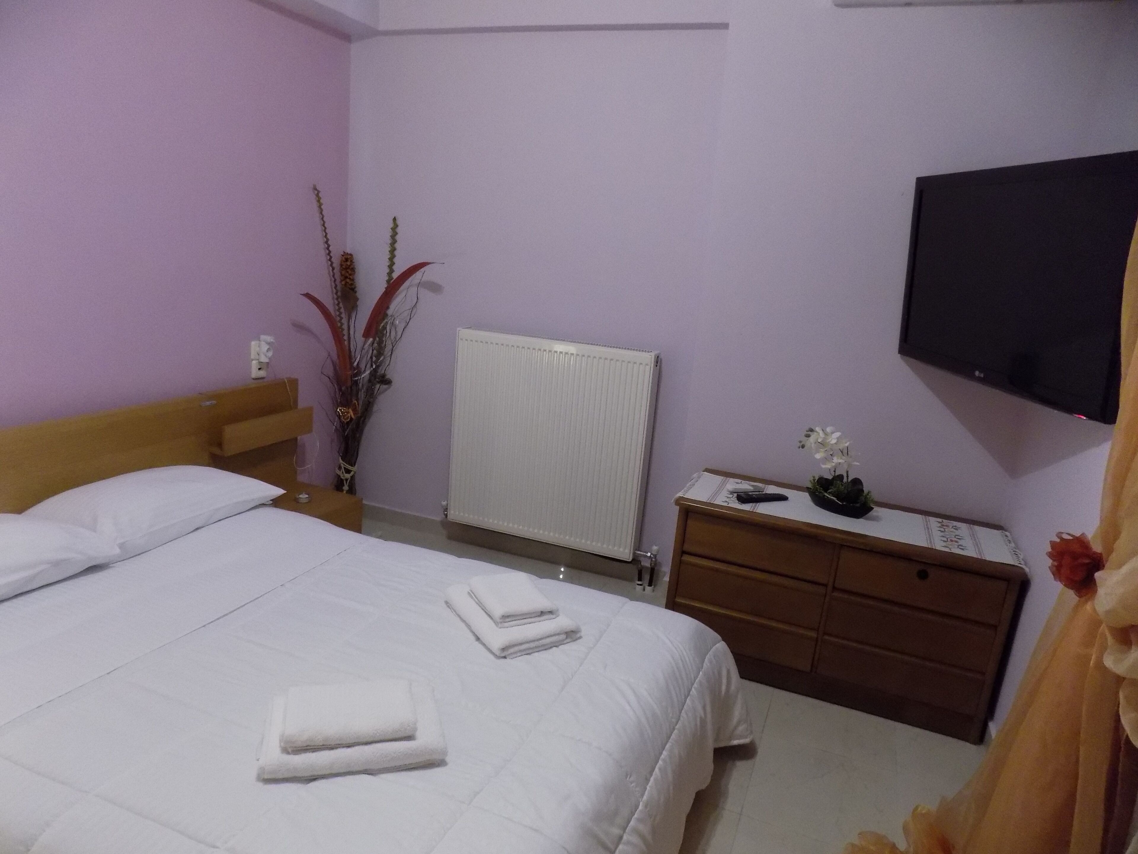 A hotel room with a bed, a bedside table, a dresser, a radiator, and a flat-screen TV mounted on the wall.