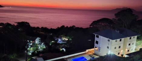 A coastal property with a swimming pool, a multi-story building, and a view of the sea at sunset.
