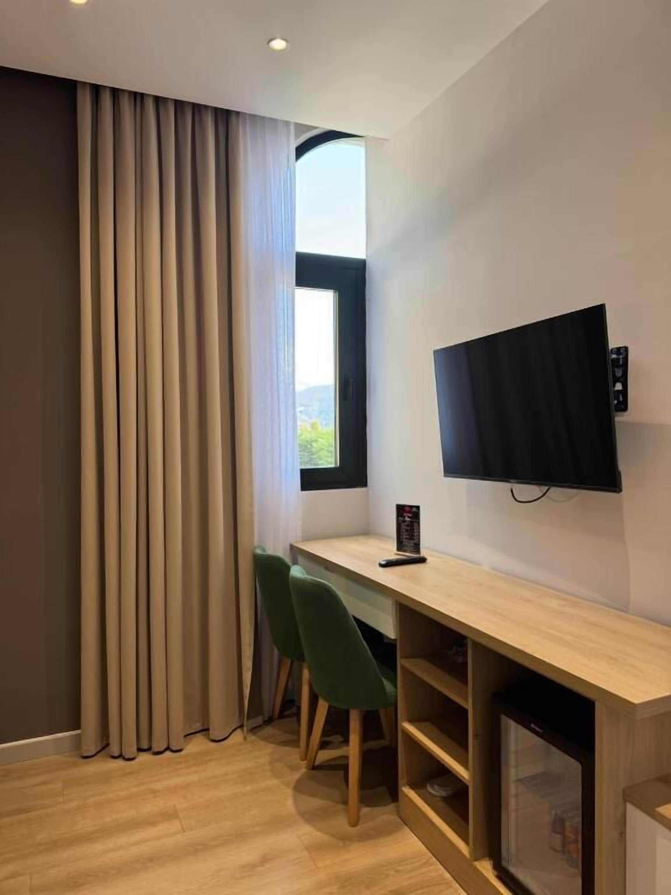 Una habitación moderna con un escritorio de madera, dos sillas verdes, un televisor montado en la pared y una ventana con cortinas.