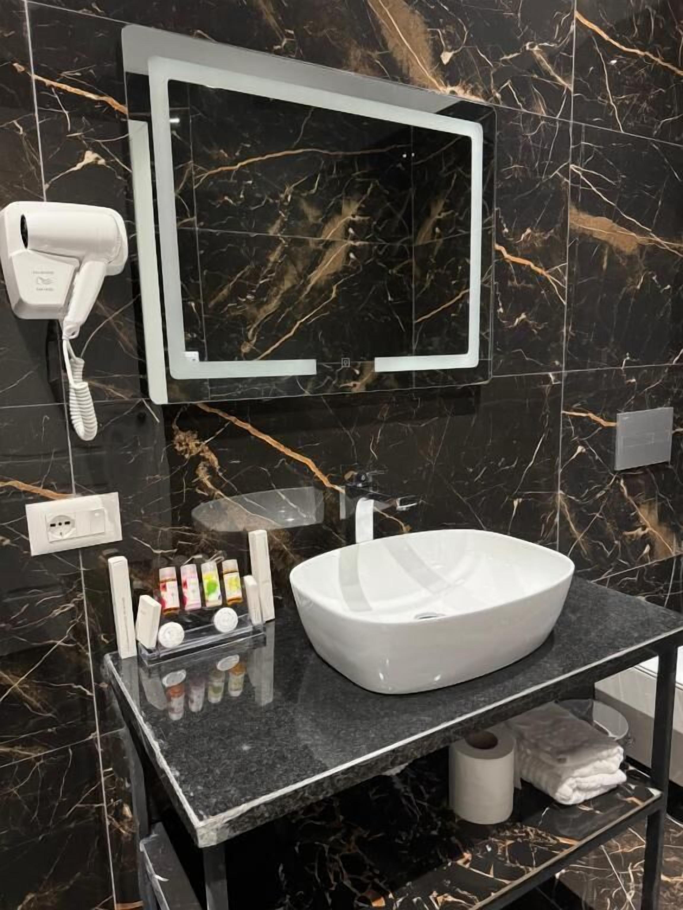 A modern bathroom with a black marble backsplash, a white sink, and a mirror above it. There is a hairdryer on the wall and a shelf with toiletries.