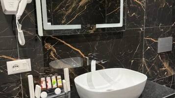 A modern bathroom with a black marble backsplash, a white sink, and a mirror above it. There is a hairdryer on the wall and a shelf with toiletries.
