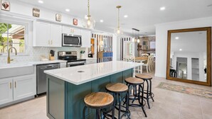 Una cocina moderna con armarios blancos, una isla grande con taburetes, electrodomésticos de acero inoxidable e iluminación colgante.