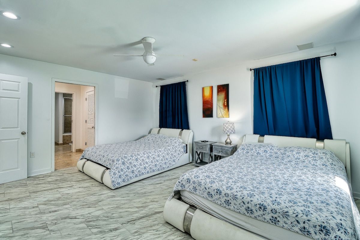 Habitación de hotel con dos camas, un ventilador de techo, un escritorio con lámpara y obras de arte en la pared.