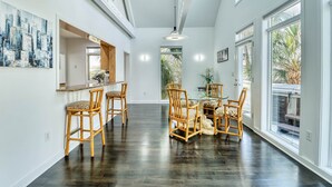 A spacious hallway with dark wooden floors, a dining area with wicker chairs and a table, and large windows offering views of greenery. - Wow By The Sea (North Myrtle Beach)