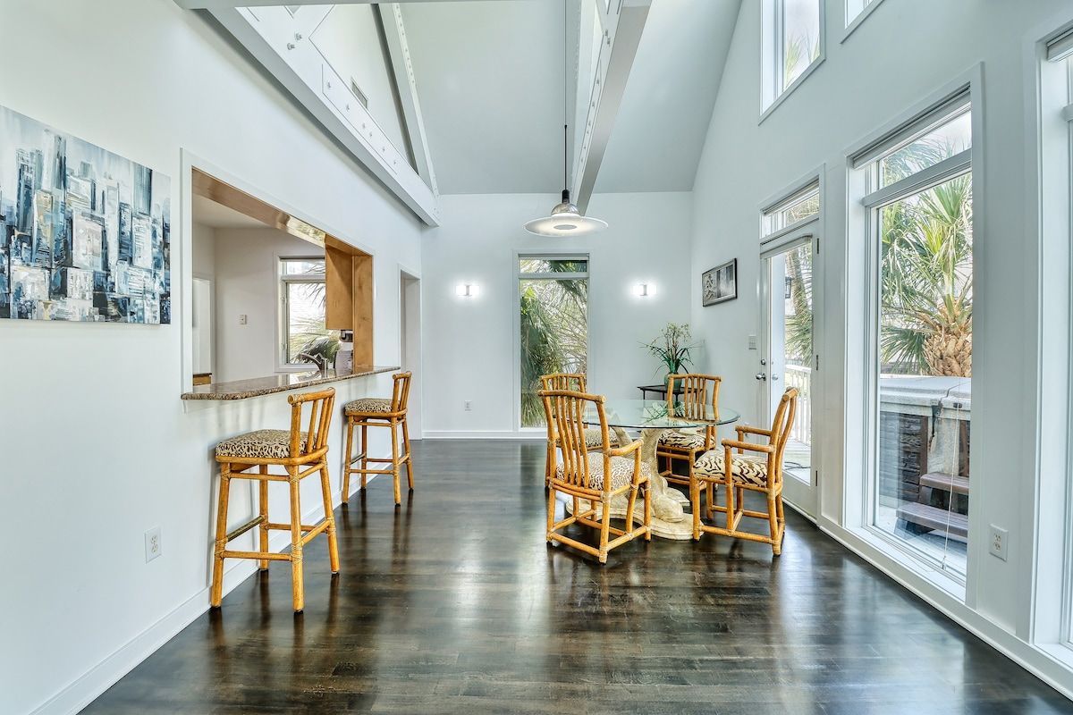 A spacious interior with a dark wooden floor, a dining area with wicker chairs, and large windows providing natural light.