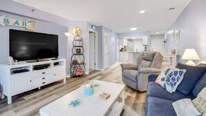 A living room with a TV, a bookshelf, a chair, a sofa, and a coffee table. - Sunset at Sassy's 2 (North Myrtle Beach)