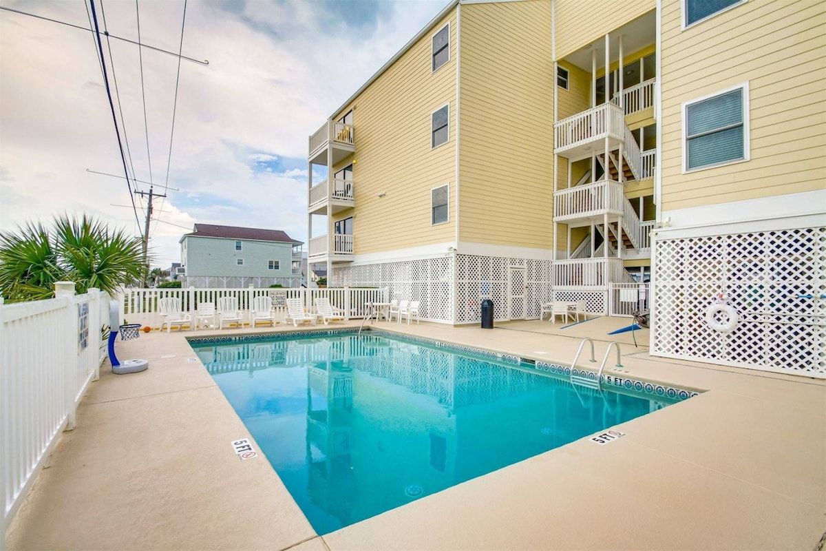 A swimming pool area with a pool, white fences, and a building with balconies.