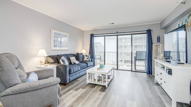 A modern living room with a grey sofa, a white coffee table, and a grey armchair. There is a white cabinet with a TV and a large window with curtains. - Sunset at Sassy's 2 (North Myrtle Beach)