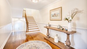 A hallway with a wooden staircase, a white door, a framed picture, a wooden console table, and a vase with dried branches. - Seaside Magnolia (North Myrtle Beach)