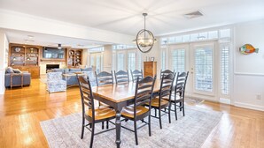 A spacious dining area with a wooden table and chairs, a chandelier, and a view through large windows. - Seaside Magnolia (North Myrtle Beach)