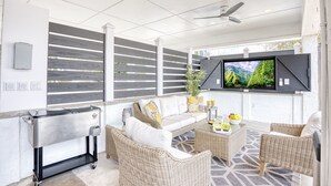 A modern outdoor living area with a sofa, wicker chairs, a coffee table, a TV, and a grill. - Seaside Magnolia (North Myrtle Beach)