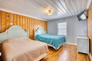 A room with two beds, wooden paneling on the walls, a ceiling fan, a television mounted on the wall, and a window with blinds.