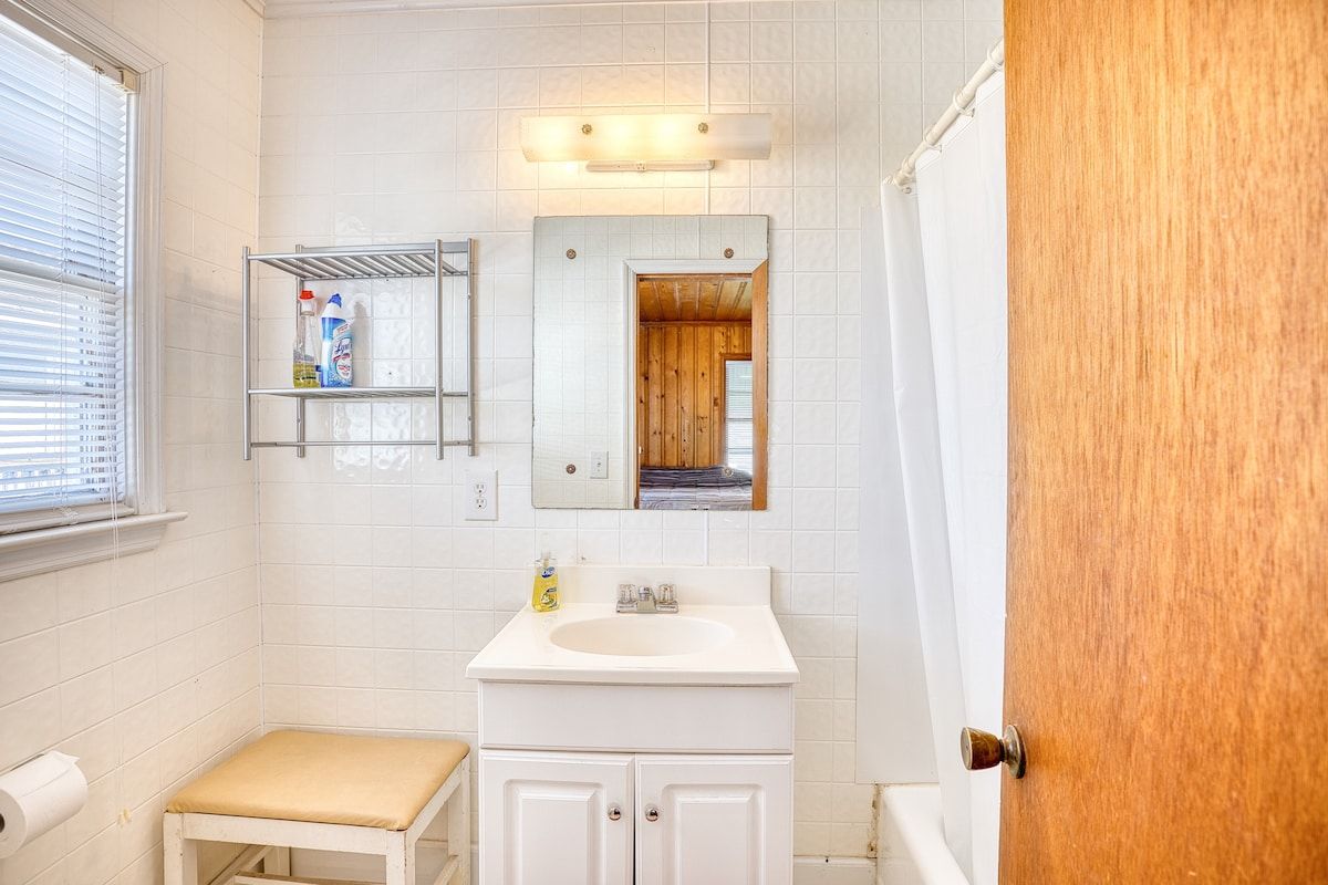 A bathroom with a white sink, a mirror, a shelf with toiletries, and a wooden door.