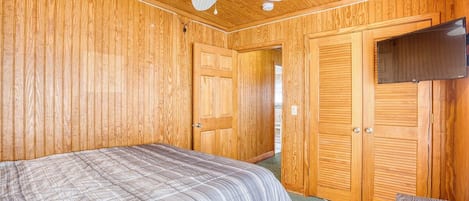 A bedroom with a bed, wooden walls, a ceiling fan, and a TV mounted on the wall.