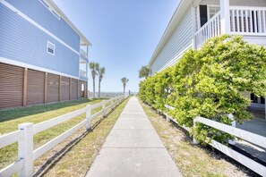 A pathway flanked by white fences and lush greenery, leading to a building with a balcony.
