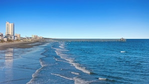 A coastal view with a pier extending into the sea, buildings along the shore, and clear skies.