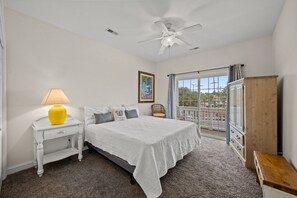 A bedroom with a bed, a nightstand, a lamp, a ceiling fan, a window with curtains, and a view of the outside.