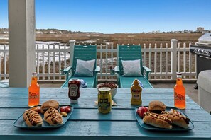 Een tafel met hotdogs, toppings en drankjes op een terras met uitzicht op een strand en een witte schutting.