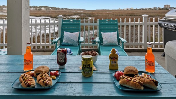 Et bord med hotdogs, tilbehør og drikkevarer på en terrasse med udsigt over en strand.
