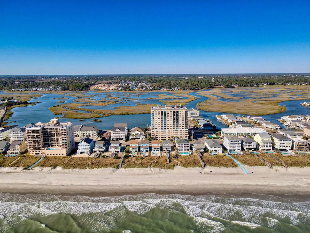 A coastal hotel with multiple buildings, beachfront access, and a marshy landscape in the background.