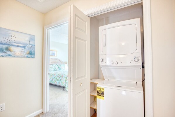 A laundry room with a washing machine and dryer, a door leading to a bedroom with a bed, and a painting on the wall.