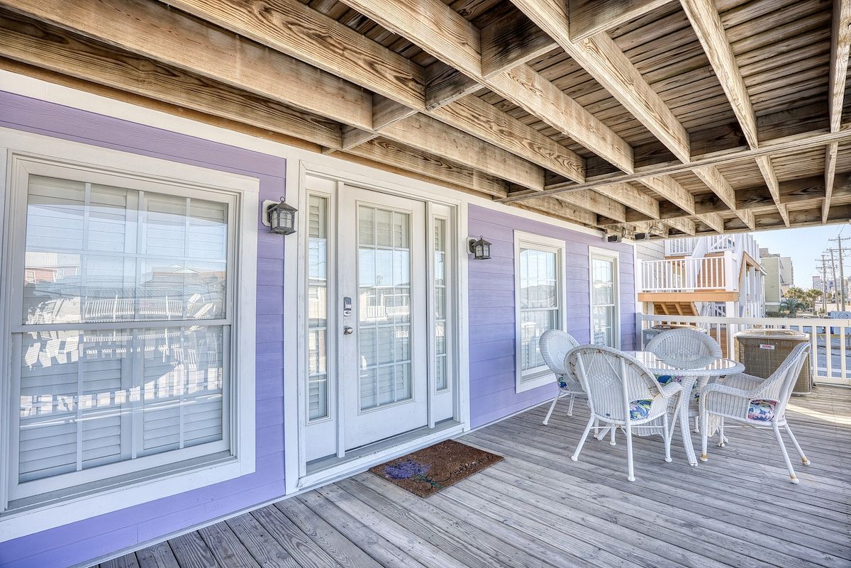 Une véranda couverte avec un plafond en bois, des meubles en osier blancs et un mur extérieur de couleur violette.