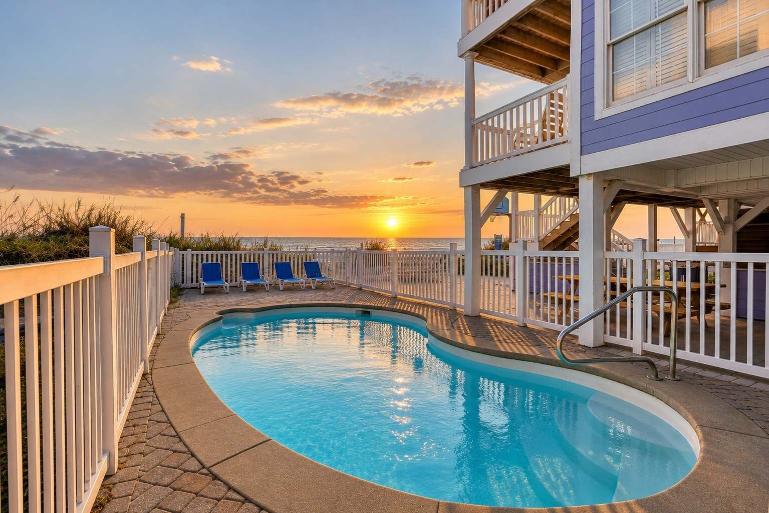 A swimming pool with a view of the sunset, a white fence, and blue lounge chairs.