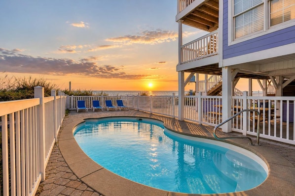 A swimming pool with a view of the sunset, a white fence, and blue lounge chairs.