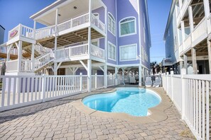 Un área de piscina con una cerca blanca, un edificio de varios niveles con balcones y un cielo azul despejado.