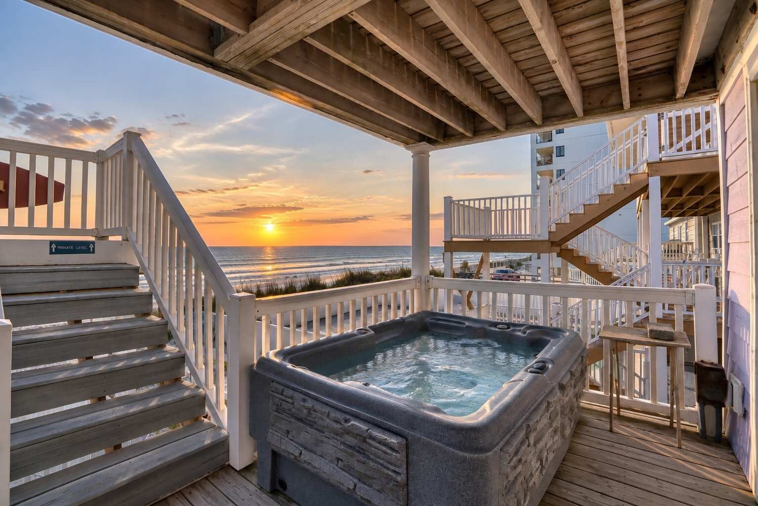 A wooden deck with a hot tub, a staircase, and a view of the ocean at sunset.