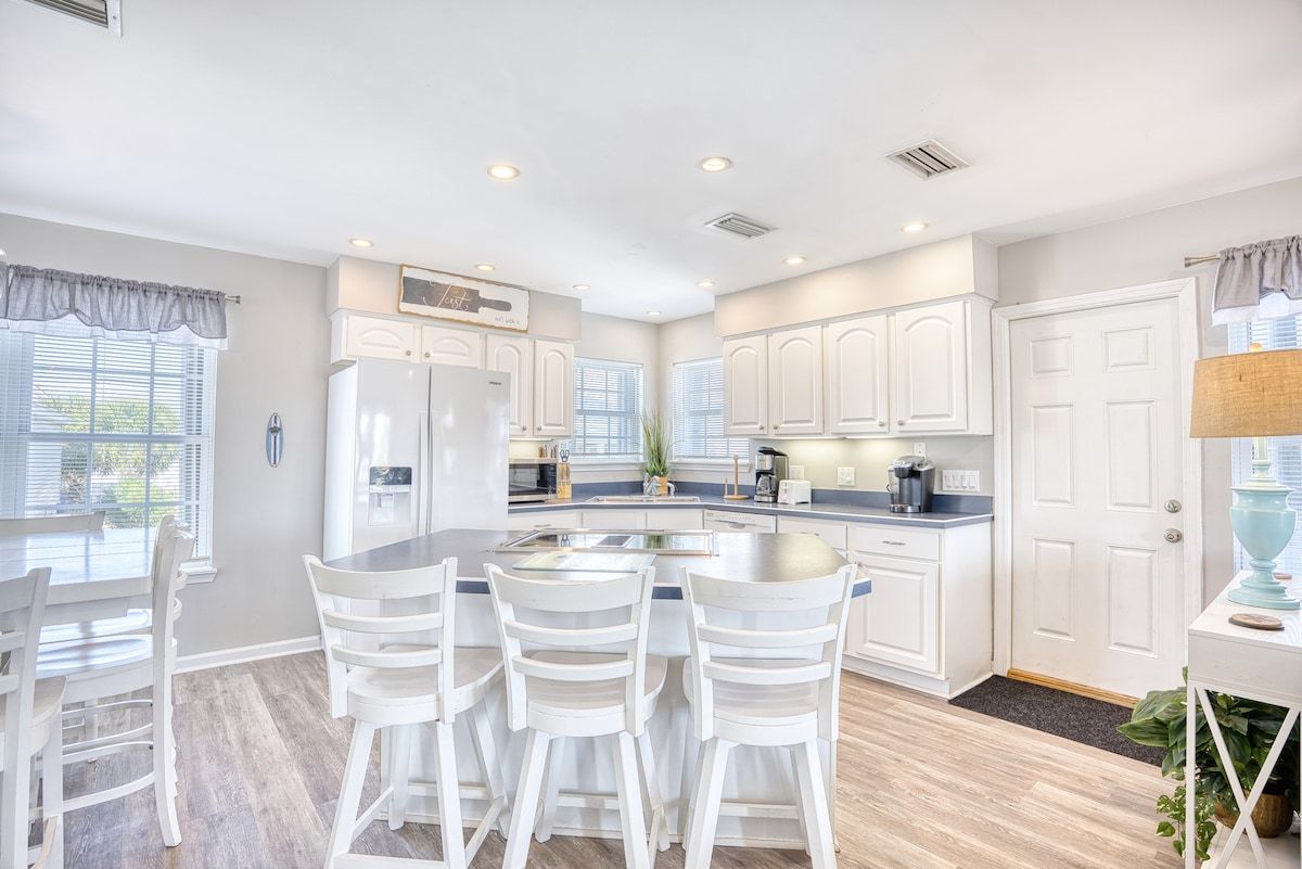 A modern kitchen with white cabinetry, a central island with seating, and a dining area with white chairs.