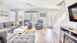 A spacious living room with a grey sectional sofa, a blue armchair, and a white coffee table. There is a ceiling fan, a TV mounted on the wall, and a staircase leading to another level. - 5 O'Clock Poolside (North Myrtle Beach)